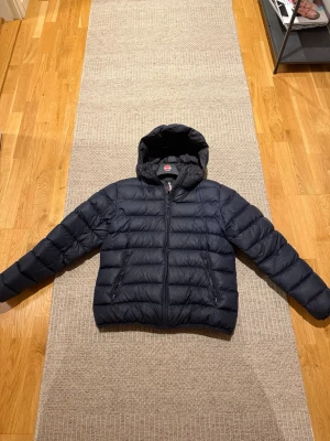 Mörkblå pufferjacka från Colmar - Säljer en mörkblå pufferjacka från Colmar med huva och dragkedja framtill. Jackan har quiltad design, två fickor med dragkedja och en rund Colmar-logga på ärmen. Perfekt för kyliga dagar och riktigt snygg till streetwear-stilen. C:et på colmar märket är borta! Den är 170cm! Pris går att diskuteras!