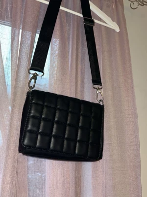 Svart väska - Säljer en svart väska med bred justerbar rem och rutmönstrad framsida. Köpte den på Ullared för några år sedan men den har knappt används därav finns inga defekter.❤️