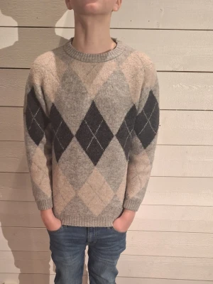 Gråmelerad stickad tröja med rutor - Säljer en gråmelerad stickad tröja med klassiskt argylemönster i svart och beige på framsidan och ärmarna. Tröjan har rund halsringning, ribbade muddar och lång ärm. Perfekt för dig som gillar retrovibbar och vill ha något riktigt mysigt till hösten.