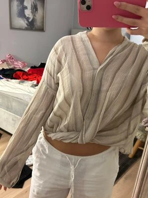 Beige randig skjorta med knyt - Luftig beige skjorta med tunna vertikala ränder i olika nyanser och subtilt glittriga trådar. Skjortan har v-ringad hals, knappar framtill och är knuten nertill för en chill vibe. Långärmad och i ett lätt, transparent material som känns perfekt för varmare dagar.