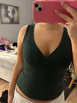 Mörkgrön omlott linnetopp - Snygg mörkgrön ärmlös linnetopp med omlott och djup v-ringning. Toppen är figurnära och har en enkel, stilren look som passar perfekt till jeans eller kjol. Mjuk och stretchig känsla, perfekt för varmare dagar.