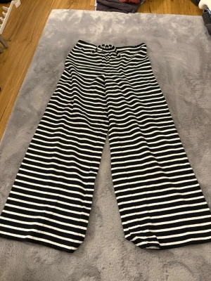 Svarta och vita randiga mjukisbyxor - Randiga mjukisbyxor från XOXO Friends i svart och vitt. Byxorna har breda ben, resår i midjan och snörning framtill. Tillverkade i mjukt ribbat material som känns skönt mot huden. Perfekta för chill dagar hemma eller när du vill ha en avslappnad look.