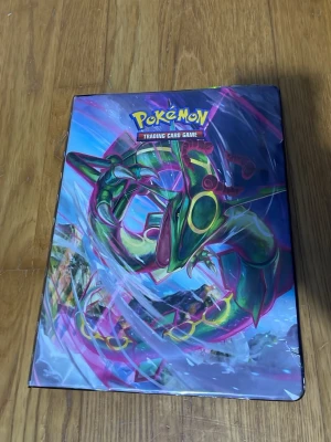 Pokémon pärm med GX och Tag Team-kort - Säljer en Pokémon pärm med färgglad Rayquaza-illustration på framsidan. Inuti finns flera sidor fyllda med GX och Tag Team-kort, många med holografisk finish. Perfekt för dig som vill boosta din samling med coola och unika kort!