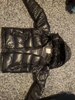 Svart Moncler Maya med huva - Säljer en svart pufferjacka från Moncler med glansig finish och huva. Jackan har två snedställda fickor med dragkedja och quiltad design. Perfekt för kalla dagar och riktigt snygg till streetwear-looken.