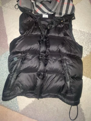 Svart dunväst från Burberry - Säljer en svart dunväst från Burberry med klassiskt rutigt foder i grått, vitt och rött. Västen har dragkedja och knappar framtill samt två fickor med dragkedja. Materialet är polyester och fodret har inslag av ull och bomull. Perfekt för lager-på-lager under höst och vinter.