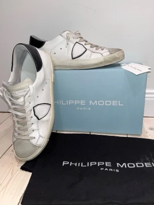 Philippe Model vita sneakers strl 42 - Säljer ett par äkta snygga Philippe Model sneakers i vitt skinn med svarta detaljer på hälen och grå mocka på tån. Klassisk låg prxs modell med mockatå och snörning. Kommer med originalkartong och dustbag. De finns slitage på sulan och lite vid plösen där blå färg, färgat av sig.  Men när man använder skorna så märker man inte slitagen. Nypris 3400 kr vilket står på kartongen. Pris kan diskuteras 🤩