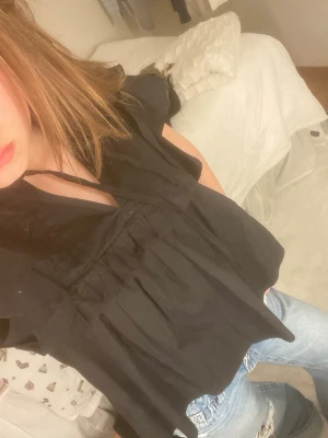 Svart blus med volangärmar - Säljer en svart blus med korta volangärmar och v-ringning med knytband. Blusen har en lös passform och är lätt rynkad under bysten för en luftig känsla. Perfekt att styla med jeans eller kjol för en trendig look.