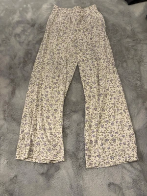 Blommiga vida byxor i bomull - Säljer ett par supermysiga, vida byxor med lila och grönt blommönster på ljus botten. Byxorna har resår i midjan och praktiska fickor på sidorna. Perfekta för chill dagar eller när du vill ha en söt och avslappnad look.