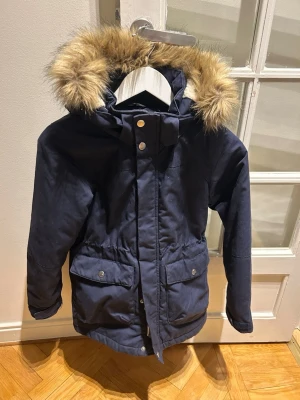 Mörkblå parkas med fuskpäls - Säljer en mörkblå parkas med huva och fuskpälsdetalj. Jackan har stora fickor framtill med tryckknappar och dragsko i midjan för snygg passform. Perfekt för kalla dagar och riktigt skön att bära.