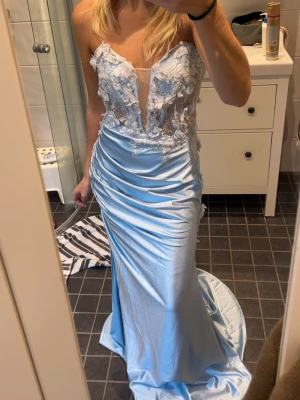 Ljusblå långklänning från Zapaka - Magisk ljusblå långklänning från Zapaka med axelbandslös design och djup v-ringning. Överdelen har spetsdetaljer, 3D-blommor och pärlor, medan kjolen är slät och glansig i satinliknande material. Perfekt för bal eller student!