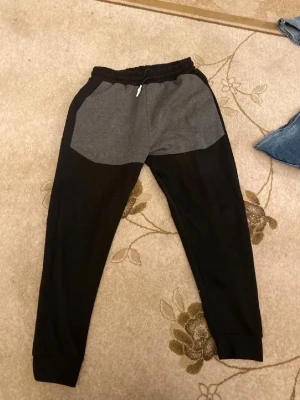 Nike mjukisbyxor svart/grå XXL - Nike mjukisbyxor i storlek XXL med svart och grå färgblock. Byxorna har elastisk midja med snörning och klassisk Nike-logga på ena sidan. Tillverkade i mjukt bomullsmaterial för skön passform och avslappnad stil.