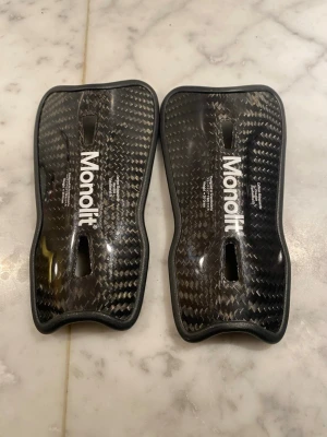 Monolit benskydd i kolfiber - Säljer ett par svarta benskydd från Monolit i kolfiber med snyggt rutmönster. De har en ergonomisk form och är superlätta, perfekta för dig som vill ha maxad rörelsefrihet på planen. Passar till fotboll och har en clean, modern look. Storlek 15cm 