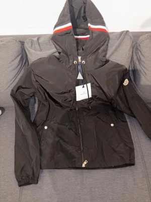 Svart vindjacka från Moncler - Snygg svart vindjacka från Moncler med huva och dragsko. Jackan har dragkedja framtill, två fickor med tryckknappar och en vit-röd-blå rand på huvan. Perfekt för dig som vill ha en stilren och sportig look.