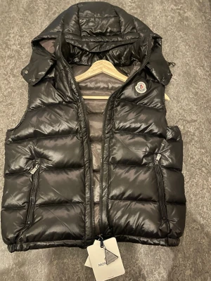 Svart dunväst från Moncler - Snygg svart dunväst från Moncler med glansig finish och avtagbar huva. Västen har två dragkedjeförsedda fickor framtill, quiltad design och klassisk Moncler-logga på bröstet. Perfekt lager-på-lager-plagg för kyligare dagar.