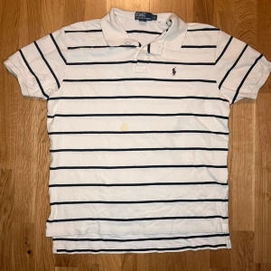 Vit randig piké från Ralph Lauren M - Vit pikétröja från Ralph Lauren med svarta horisontella ränder och klassisk krage. Tröjans size stämmer inte riktigt och sitter mer som en M. Broderad logga i vinrött på bröstet. Tillverkad i bomull och har korta ärmar. Perfekt för en clean och sportig stil.