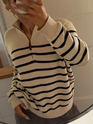 Randig beige half zip tröja - Säljer en beige stickad tröja med svarta ränder och half zip-dragkedja. Tröjan har hög krage, ribbade muddar och en relaxed, oversized passform. Perfekt för dig som gillar en chill och stilren look.