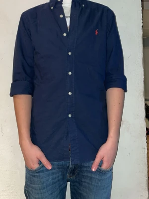 Marinblå skjorta från Ralph Lauren - Klassisk marinblå skjorta från Ralph Lauren med knappslå och button-down krage. Skjortan har långa ärmar som är uppvikta och den ikoniska röda loggan broderad på bröstet. Tillverkad i mjuk bomull för en skön känsla.