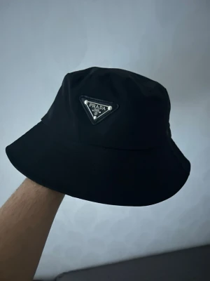 Svart bucket hat från Prada - Säljer en svart bucket hat från Prada med klassisk trekantslogga framtill. Hatten är i ett slätt, syntetiskt material och har en enkel, stilren design. Perfekt accessoar för att lyfta din outfit.