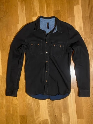 Nudie Jeans Jeansskjorta  - Säljer en svart långärmad skjorta från Nudie Jeans med tryckknappar i metall och två bröstfickor. Skjortan har klassisk western-inspirerad design och är gjord i mjuk bomull. Insidan av kragen är fodrad med blått tyg för en snygg kontrast. Ifall ni vill ha föer bilder eller om ni har frågor skriv!