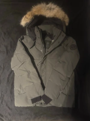Grå dunjacka från Canada Goose - Säljer en grå dunjacka från Canada Goose med pälsdetalj på huvan och klassisk patch på ärmen. Jackan har dragkedja och knappar framtill, långa ärmar och en riktigt varm och skön känsla. Perfekt för kalla vinterdagar och snygg till alla outfits.
