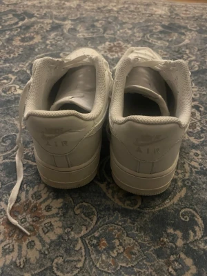 Nike Air Force 1 vita sneakers - Klassiska Nike Air Force 1 sneakers i helvitt skinn med rund tå och snörning. Snygg och tidlös modell med perforerade detaljer på tån och den ikoniska swoosh-loggan på sidan. Passar perfekt till streetwear och casual outfits.