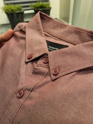 Rosa skjorta Reserved Slim Fit M - Säljer en rosa skjorta från Reserved i slim fit-modell. Skjortan har långa ärmar, svarta armbågslappar och matchande rosa knappar. Klassisk krage och mjukt bomullstyg gör den både snygg och bekväm. Perfekt för dig som gillar stilrena detaljer.