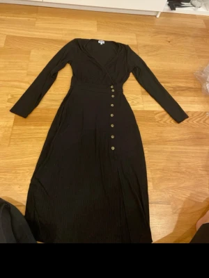 Svart långklänning från Bubbleroom - Svart långklänning från Bubbleroom med omlott-topp, v-ringning och långa ärmar. Klänningen har ribbad struktur, dekorativa knappar längs framsidan och en liten slits. Perfekt för dig som gillar stilrena och bekväma plagg.