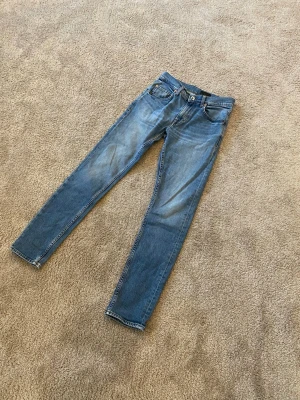 Blå jeans från Tiger of Sweden - Säljer ett par klassiska blå jeans från Tiger of Sweden. De har en rak passform med fem fickor, snygga kontrastsömmar och läderpatch bak i midjan. Jeansen är i ett mjukt bomullsmaterial och har en tidlös look som funkar till allt.