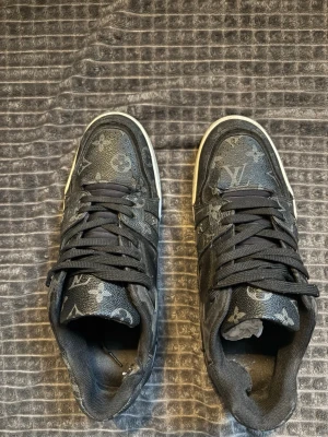 Louis Vuitton svarta sneakers monogram - Säljer ett par svarta Louis Vuitton sneakers med klassiskt monogrammönster över hela skon. Skorna har vita sulor med LV-detaljer på sidan och undertill. Tillverkade i skinn och textil med svarta snören och rund tå. Riktigt snygga och unika statement sneakers.