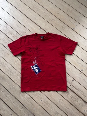 Röd Vintage Y2K Quiksilver t-shirt med tryck - Snygg röd t-shirt från Quiksilver med grafiskt tryck i svart, vitt och marinblått på framsidan och mindre tryck på ryggen. Två fläckar, se sista bilder. Annars i fint skick. Inget slitage på tryck.  Lapparna på tröjan säger olika storlek, XL och L. Men tröjan passar som M.   Kontakta mig ifall mått önskas.