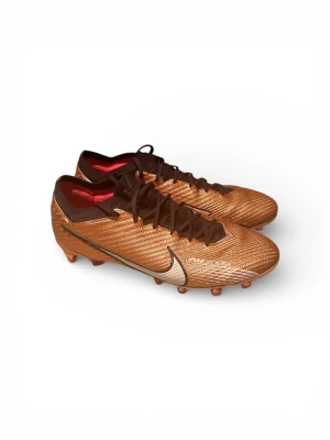 Nike fotbollsskor i koppar och brunt - Säljer ett par riktigt snygga Nike fotbollsskor i kopparfärg med bruna detaljer och snörning. Skorna har en skinande metallic finish, låg profil och mönstrad ovandel i syntetmaterial. Sulan har fasta dobbar och coola röda inslag undertill. Perfekta för dig som vill sticka ut på planen. Pris kan diskuteras vid snabb affär!