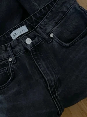 Svarta raka jeans från Zara - Säljer ett par svarta jeans från Zara med raka ben och rå kant nertill. Klassisk femficksmodell med silverfärgade knappar och detaljer. Jeansen har medel midja och är tillverkade i ett robust bomullstyg.