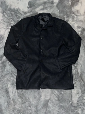 Svart ullrock med knappar - Snygg svart rock i ullmix med klassisk rak passform och ståkrage. Jackan har helknäppning med stora knappar framtill och två diskreta sidofickor. Fodrad insida i blankt tyg för extra komfort. Perfekt för dig som vill ha en stilren och enkel look.