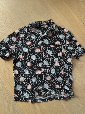 Rick and Morty skjorta från H&M - Kortärmad skjorta från H&M med Rick and Morty-tryck över hela plagget. Skjortan är svart med färgglada ansikten och vita mönster, har klassisk krage och knäppning framtill. Perfekt för dig som vill sticka ut med en unik och rolig stil.