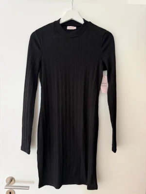 Svart ribbad klänning från Nelly - Säljer en svart ribbad kortklänning från Nelly med lång ärm och rund halsringning. Klänningen är i mjukt stickat material och har en enkel, stilren design som är lätt att matcha med olika accessoarer. Lite genomskinlig. 