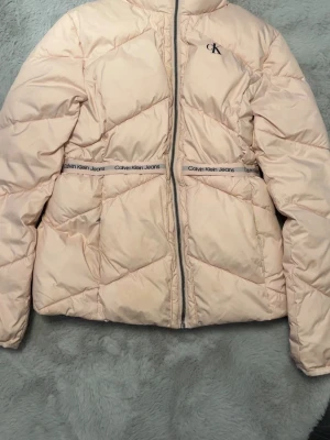 Beige pufferjacka från Calvin Klein Jeans - Säljer en beige/rosa pufferjacka från Calvin Klein Jeans med dragkedja framtill och logga på bröstet. Jackan har långa ärmar, quiltad design och band med Calvin Klein Jeans-tryck i midjan. Perfekt för kalla dagar och riktigt snygg till alla outfits. Äkta såklart och priset kan diskuteras