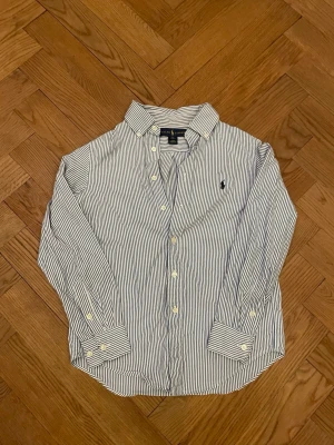 Randig skjorta från Ralph Lauren - Klassisk skjorta från Ralph Lauren i vit och blå med tunna ränder. Skjortan har lång ärm, knappar framtill och broderad logga på bröstet. Tillverkad i mjuk bomull och har en snygg krage. Perfekt för dig som gillar preppy stil.