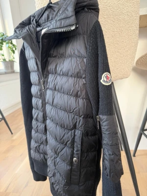 Svart Moncler pufferjacka med huva - Säljer en svart pufferjacka från Moncler med stickade ärmar och vadderad kropp. Jackan har huva, dragkedja framtill, fickor med tryckknapp och en extra ficka med dragkedja på ärmen. Klassisk Moncler-logga på armen och ribbade muddar för extra komfort.