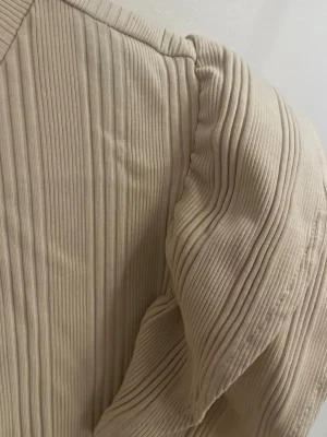 Beige ribbad topp med volangärm Gina Tricot - Säljer en beige ribbad topp från Gina Tricot i storlek 158/164. Toppen har korta volangärmar och rund halsringning. Materialet är mjukt och stretchigt, perfekt för en chill och snygg look. Passar till allt och är superenkel att styla.