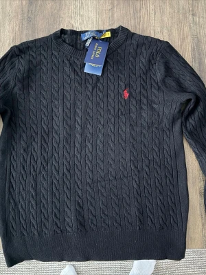 Svart kabelstickad tröja Polo Ralph Lauren - Kabelstickad svart tröja från Polo Ralph Lauren med klassisk röd logga broderad på bröstet. Rund halsringning och ribbade muddar. Perfekt till lager på lager och riktigt snygg till jeans eller kjol.