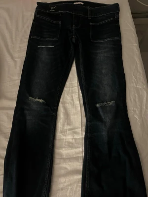 Mörkblå bootcut jeans med slitningar - Säljer ett par mörkblå jeans med bootcut passform och snygga slitningar vid knäna. Jeansen har klassiska fem fickor, kontrastsömmar och är tillverkade i ett robust denimtyg. Perfekta för dig som gillar en avslappnad men trendig look. 🙌