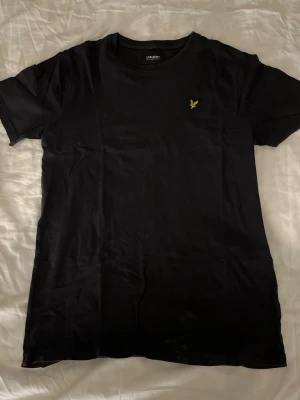 Svart t-shirt från Lyle & Scott - En clean svart t-shirt från Lyle & Scott med klassisk rund hals och det ikoniska gula fågel-loggan broderad på bröstet. Tillverkad i mjuk bomull och har korta ärmar. Står junior men skulle även funka till personer med storlek S-M