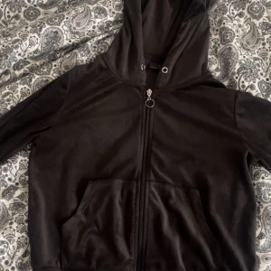 Svart velour hoodie med dragkedja - Svart hoodie i mjuk velour med huva och silverfärgad dragkedja med ringdetalj. Tyvärr har hoodien inga snören. Hoodien har långa ärmar, känguruficka framtill och ribbade muddar. Perfekt för chill dagar och snygg att styla med jeans eller mjukisbyxor.