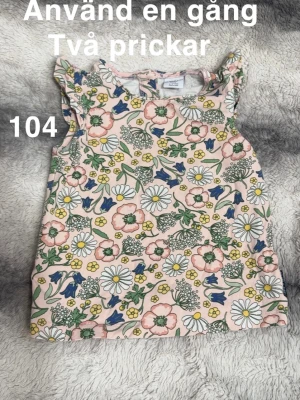 Blommig ärmlös topp med volang - Supersöt ärmlös topp med volangdetaljer på axlarna och ett färgglatt blommönster i rosa, blått, grönt, gult och vitt. Perfekt för soliga dagar och lek. 🌸 Använd en gång men fick två prickar. Storlek 104