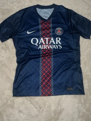 PSG matchtröja Nike blå/röd - Säljer en officiell Paris Saint-Germain matchtröja från Nike i marinblått med röd och vit geometrisk rand mitt på. Tröjan har PSG-logga på bröstet, vit Nike-swoosh och Qatar Airways-tryck. Tillverkad i lätt polyester, perfekt för fotboll eller som streetwear.