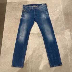 Diesel Jeans - Snygga blå jeans från Diesel, modell Belther. Jeansen har en slim-tapered passform med klassisk femficksdesign och subtila slitningar framtill. Stretchmaterial för extra komfort och snygg faded effekt på benen. Perfekt för dig som gillar en modern och avslappnad stil. Storlek 30-30 