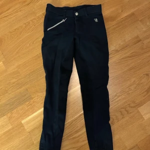 Svarta ridbyxor från Horse Life - Snygga svarta ridbyxor från Horse Life med silverfärgad dragkedja och broderad logga framtill. Byxorna har smal passform och förstärkt tyg på insidan av benen för extra grepp vid ridning. Klassisk midja och bälteshällor för att kunna använda bälte.