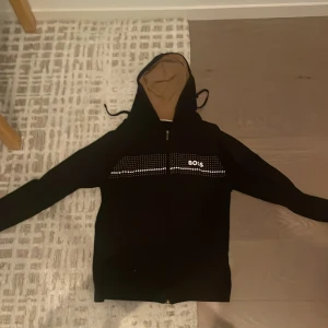 Svart hoodie med paljettdetalj från BOSS - Svart hoodie från BOSS i storlek S med snygg paljettrad över bröstet och vit logga. Tröjan har huva med snörning, dragkedja framtill och långa ärmar. Insidan av huvan är beige vilket ger en cool kontrast. Perfekt för dig som gillar streetstyle.