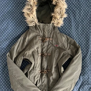 Vintage olivgrön parkajacka från Hollister - Snygg vintage olivgrön cropped parkajacka från Hollister med fluffig fuskpäls på huvan och mjukt vitt teddyfoder på insidan. Jackan har långa ärmar, tre snörningsdetaljer framtill och en broderad Hollister-logga på bröstet. Perfekt för kalla dagar och chill stil. Vintage storlek L men passar mer som M. Jackan är lite sliten runt ärmsluten men det borde inte påverka slitaget. Har bilder om det behövs. Ungefärliga mått (platta): Axel till axel: 36 cm Axel till botten: 49 cm Längs nederkanten: 