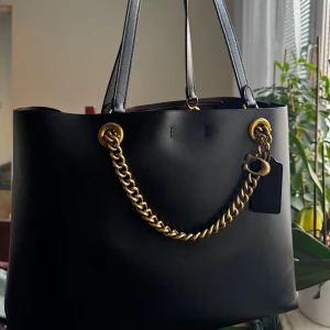 Svart handväska i skinn från Coach - Säljer min exklusiva och tidlösa Coach-väska i svart läder med guldfärgade detaljer. Väskan är rymlig, stilren och passar perfekt både till vardag och jobb. Den bärs bekvämt på axeln med dubbla remmar och har dessutom den ikoniska Coach-kedjan som extra detalj.  Skick: Nästan helt oanvänd – i mycket fint skick. Material: Slätt, högkvalitativt läder Färg: Svart med guldfärgade metall-detaljer Detaljer: Avtagbar Coach-tag, kraftig kedja, stilren design Ingår: Original dustbag från Coach